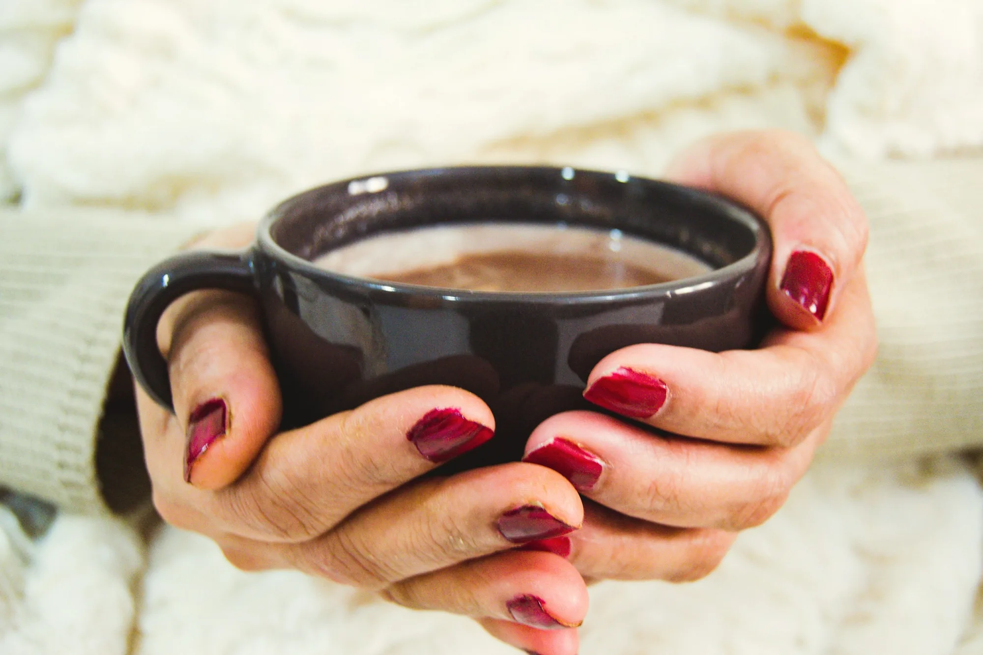 Hands with deep red nails holding a mug of hot chocolate against a cream knit sweater