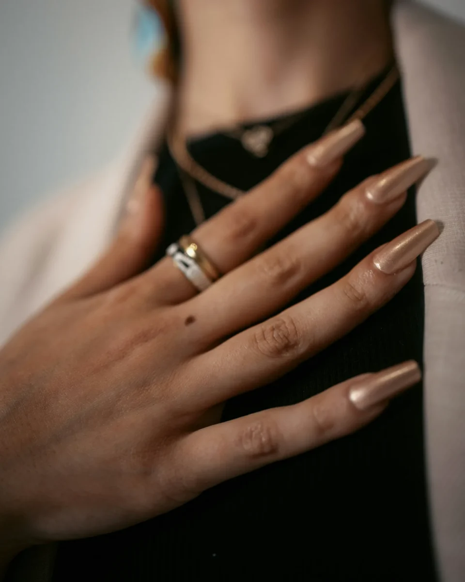 Long sculpted almond nails in nude with gold rings against a dark background