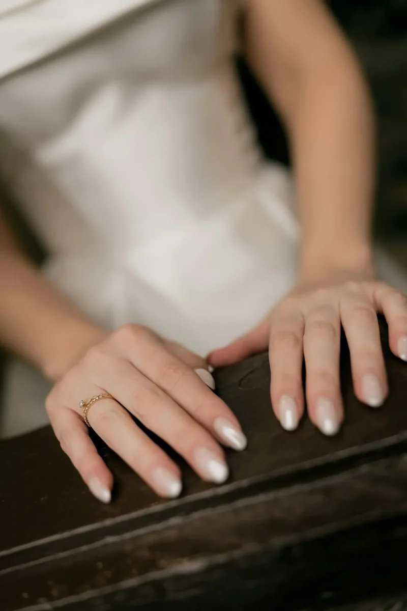 Hands with pearly nude almond nails and a fine ring resting on dark wood