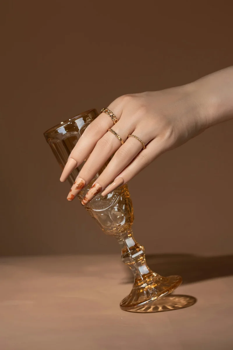 Hand with long nude almond nails holding an amber glass against a warm brown backdrop