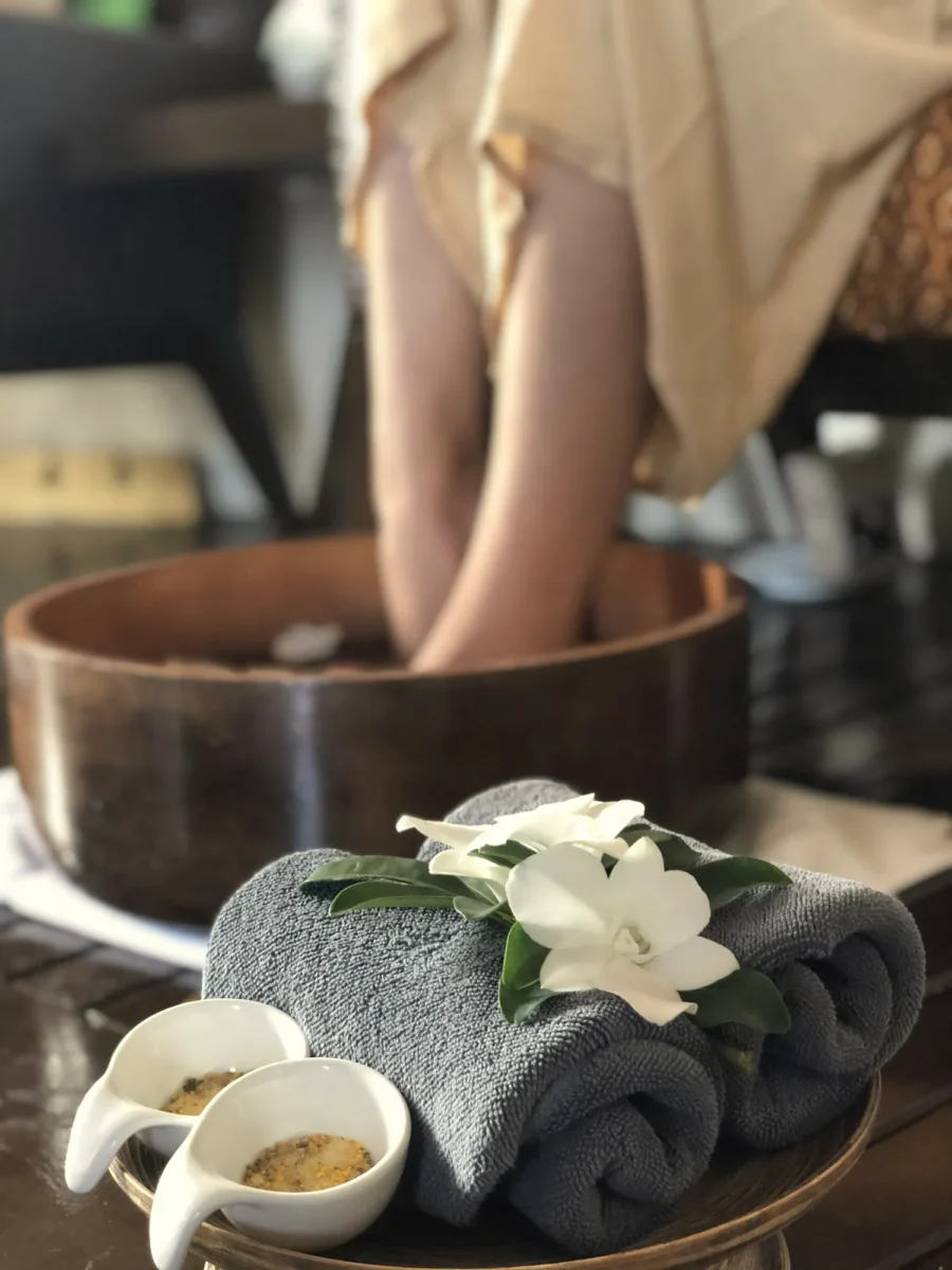 Foot soak in a wooden bowl beside rolled towels and a white flower